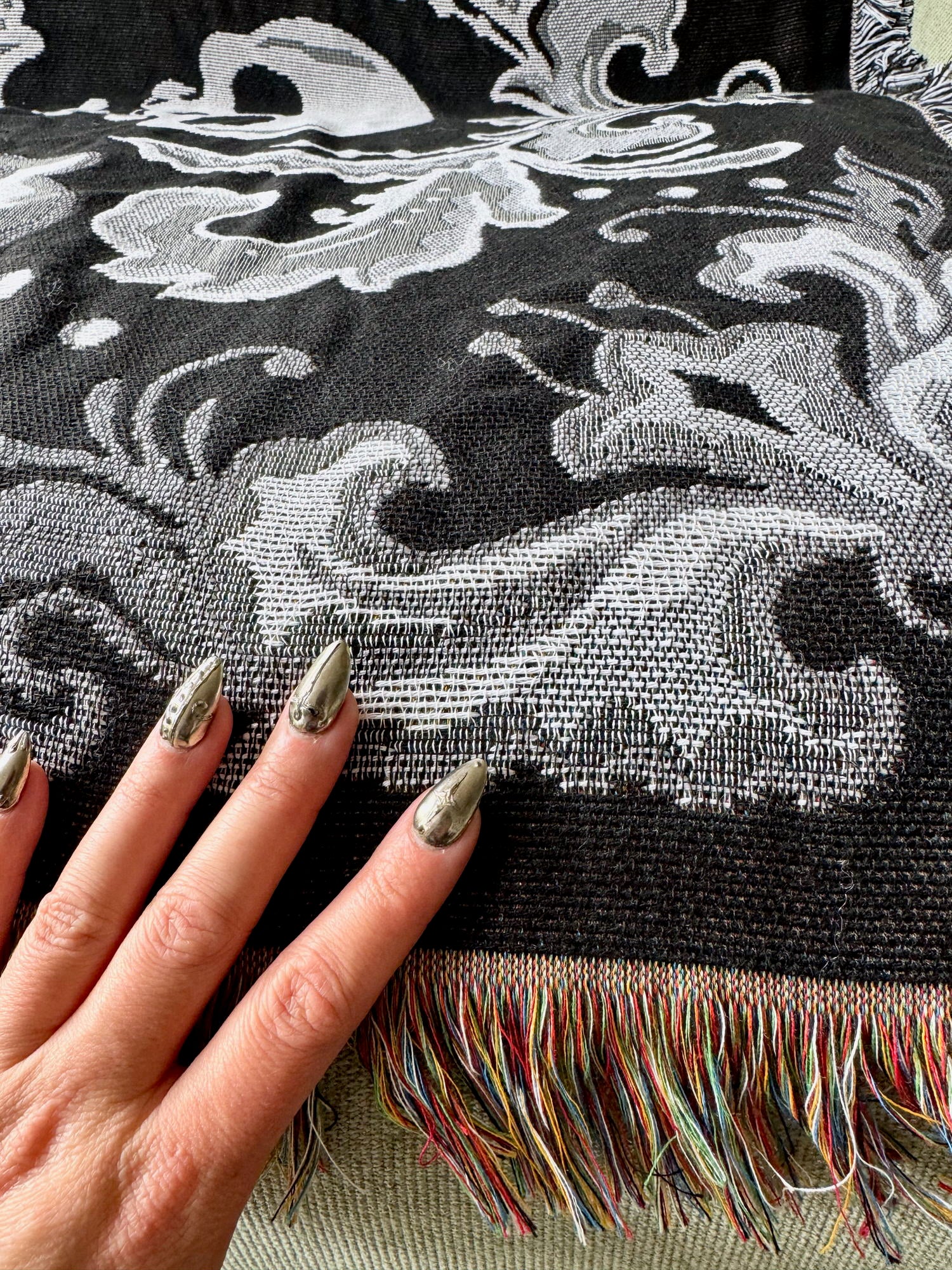 Close-up of a hand with metallic nail polish holding a black and white patterned fabric with colorful fringe.