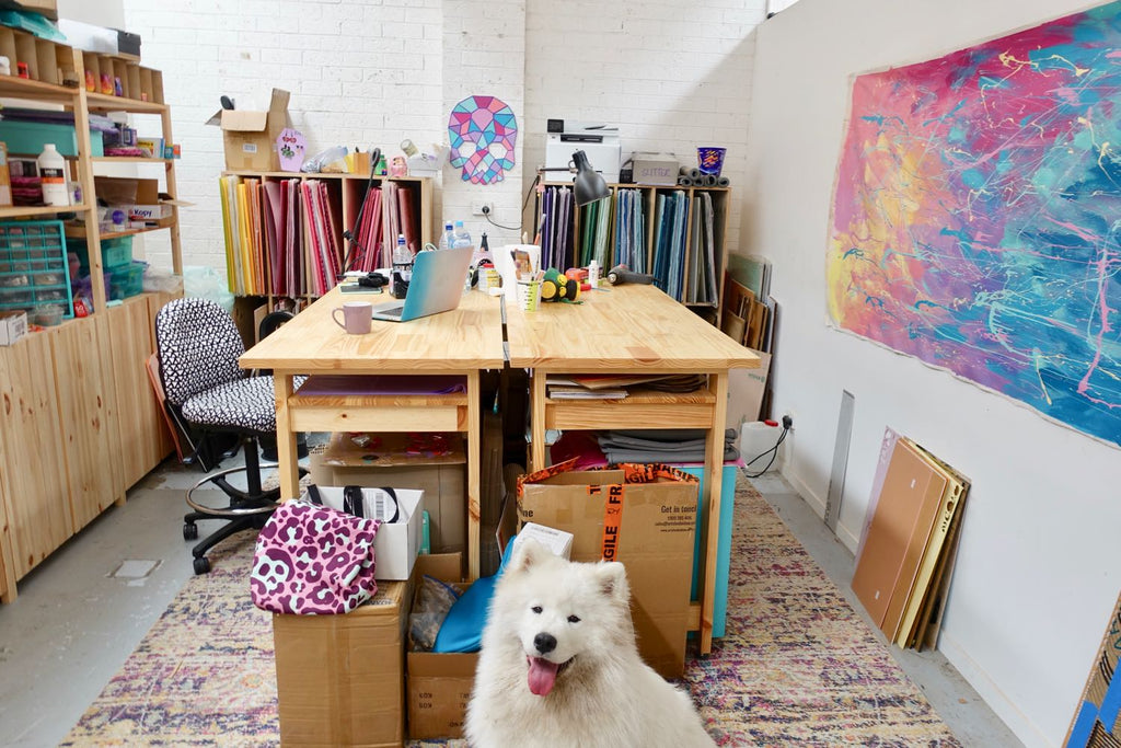 Image of a studio with pin shelving and two large workbenches, lots of clutter, and a happy fluffy white dog in the foreground