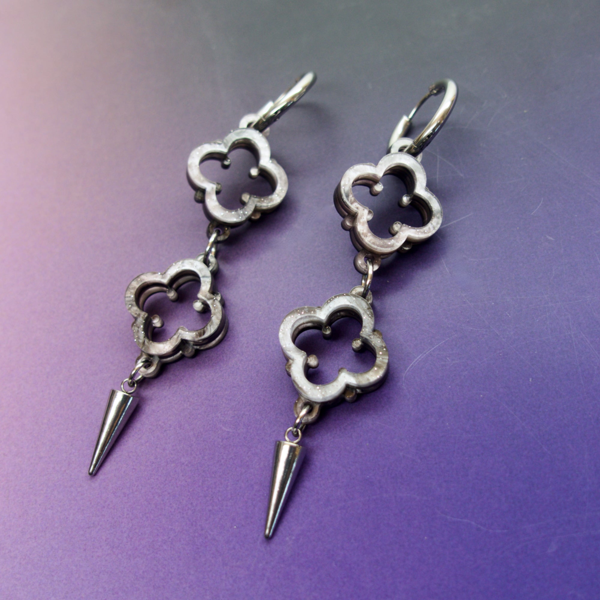 A pair of gothic-themed earrings featuring quatrefoil designs with spikes, set against a purple background.
