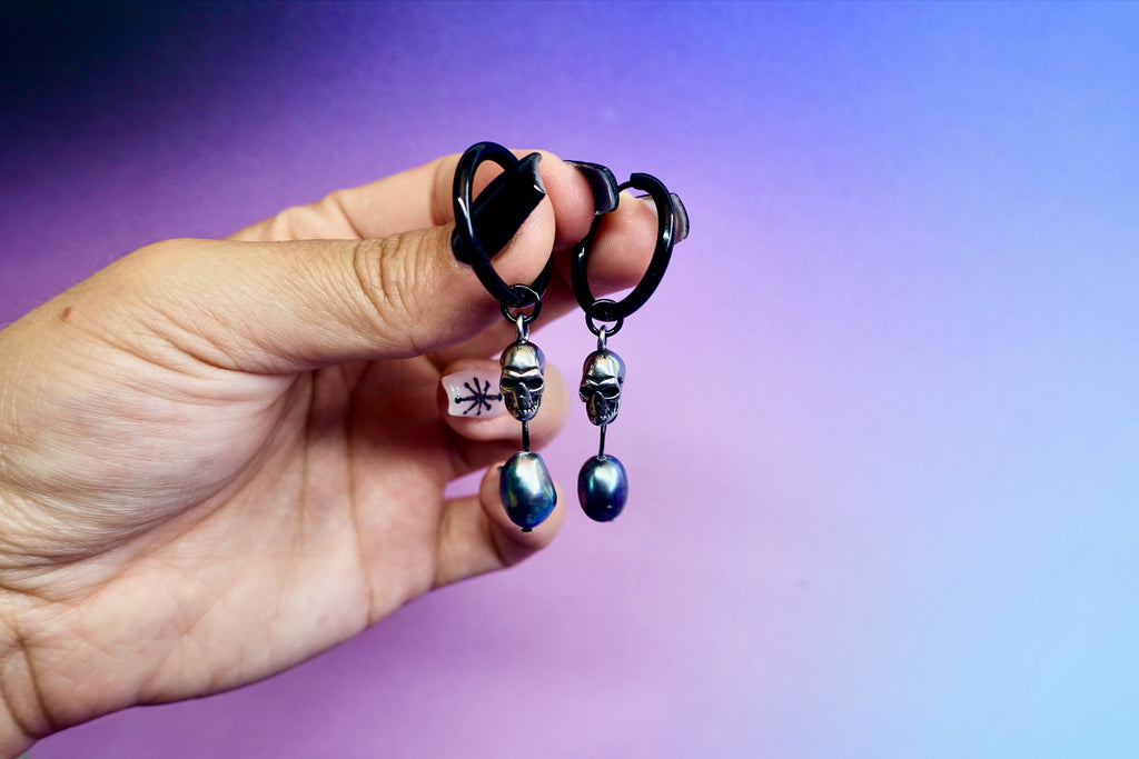 A hand holding a pair of earrings - black hoops with a steel skull and black nugget pearl hanging from them against a pastel gradient background