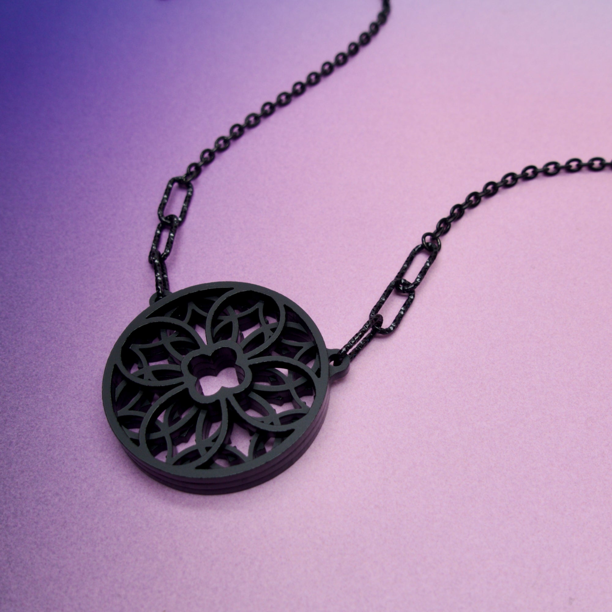 A black gothic cathedral rose window pendant necklace on a stainless steel chain against a purple background.
