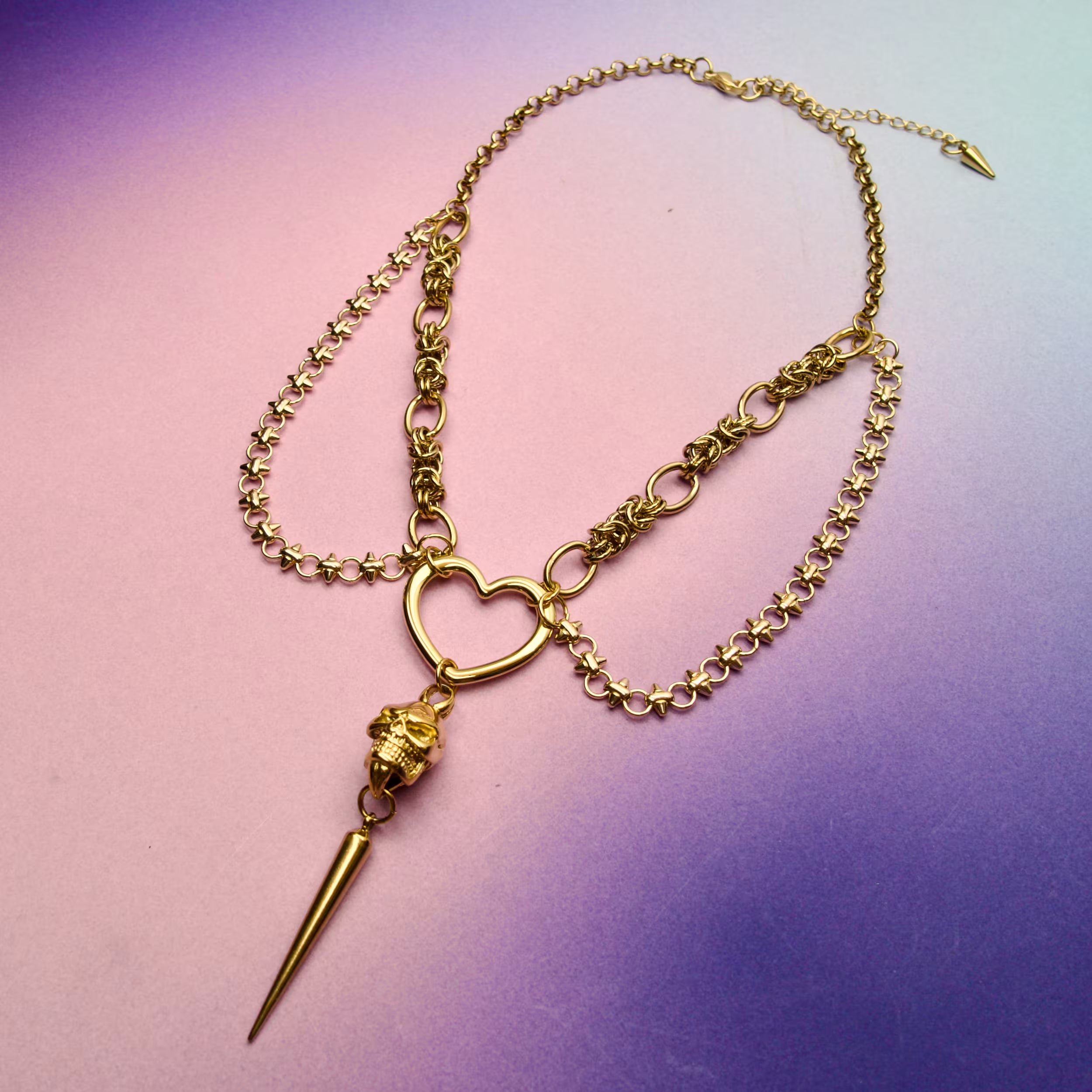 Gold gothic skull necklace with layered chains and heart centrepiece displayed on a soft gradient background.